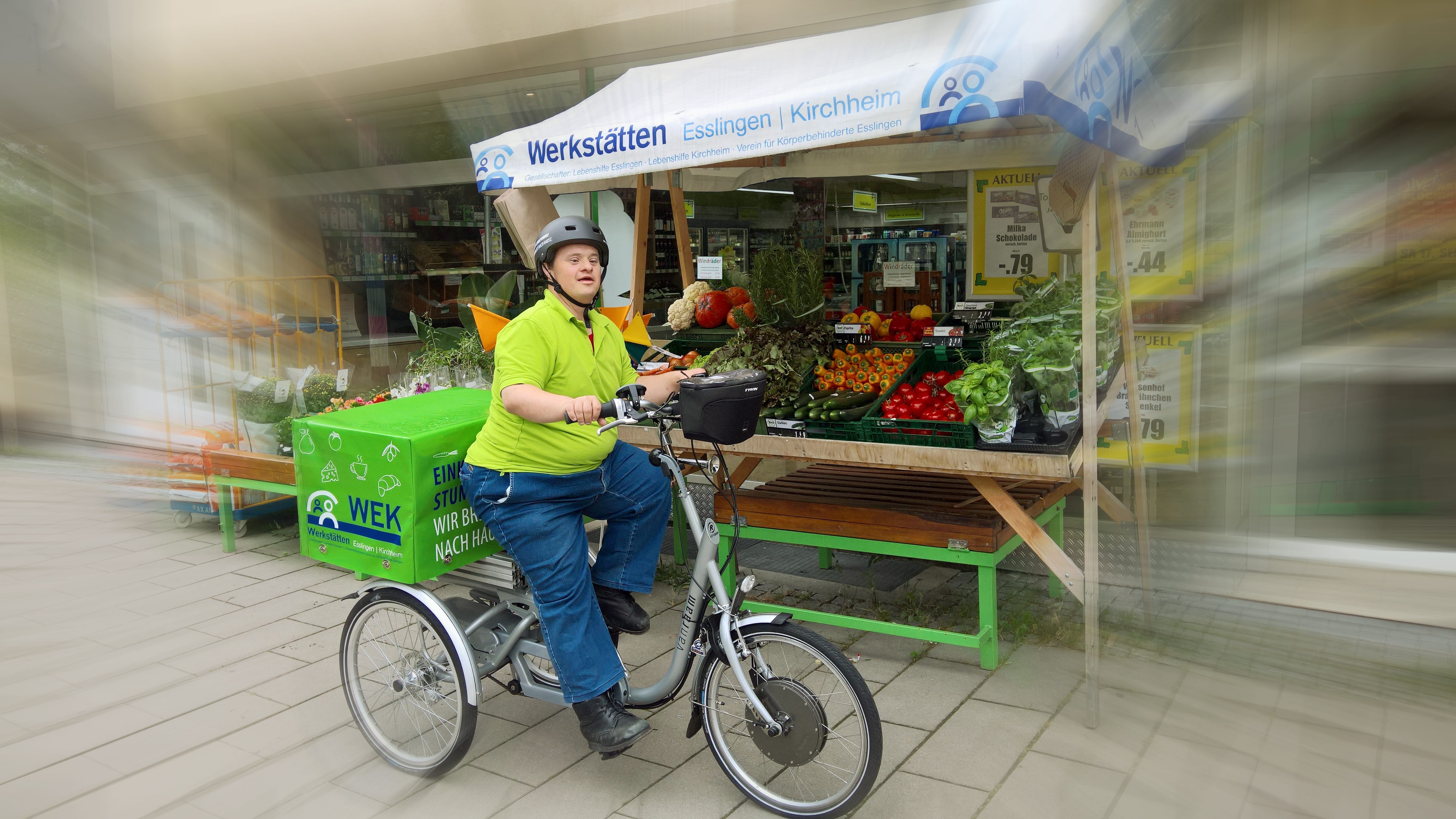 Person mit Down-Syndrom auf einem Lastenfahrrad mit Transportbox. Im Hintergrund ist ein Marktstand mit frischem Gemüse zu sehen.
